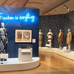 A wide gallery photo showing mannequins in dresses and a neon sign in blue that says Freedom is Everything.