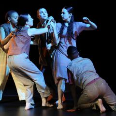 A group of five dancers huddle together on a dark stage. One dancer faces outward, looking up.