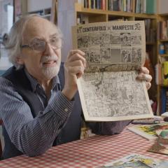 Art Spiegelman sits at a table in his home holding up a newsprint of illustration titled, Centerfold Manifesto, to the camera.