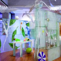 Photo of the Installation in a gallery space. Three glass walls surround a table with colorful objects. Above the walls, larger-then-life feathers suspend from the ceiling up into a skylight. 