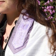 A woman wearing a purple tallit and silver Star of David necklace standing outside in front of a bush of purple flowers.