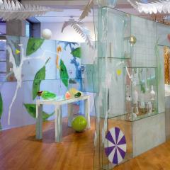 Three glass walls surround a table with colorful objects. Above the walls, larger-then-life feathers suspend from the ceiling up into a skylight.