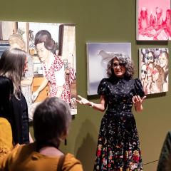 A woman artists talks to a group of people in front of her painting hung in a gallery.