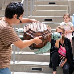A man performs with a turtle puppet in front of families in an outdoor theater.