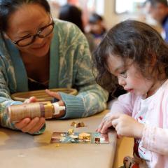 An adult helps a child on their craft project by holding the glue for them.