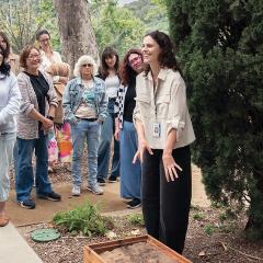 A woman speaks to a group of adults outside in a garden.