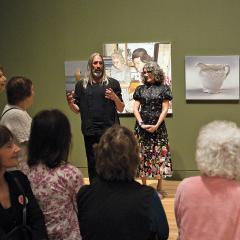 Two artist stand in front of their artwork in a gallery of paintings talking to guests.