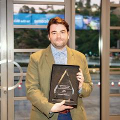 A man holds up an award smiling at the front doors of a building.
