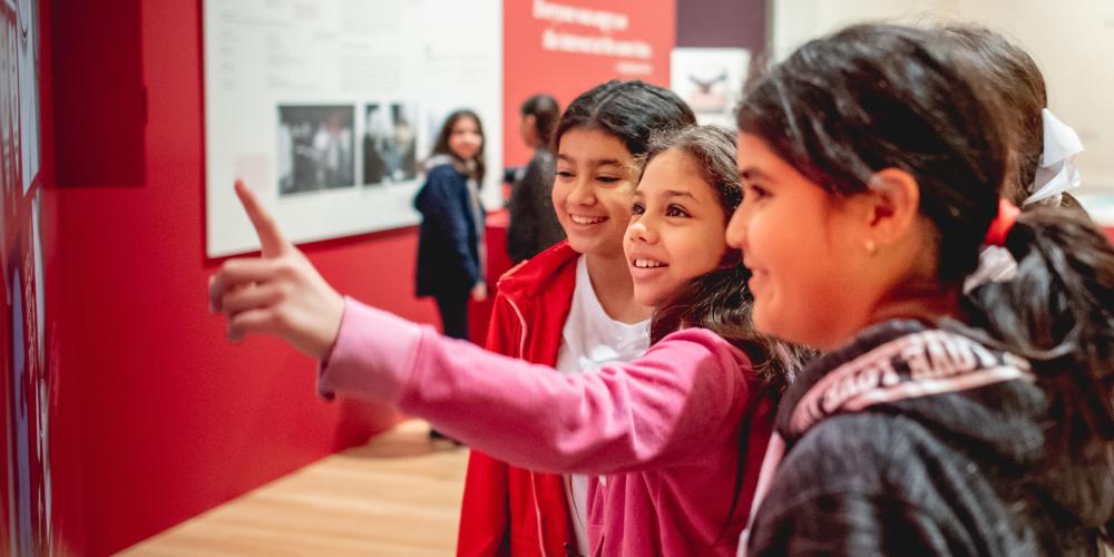 Three young girls are standing together in a red gallery looking and pointing at something out of frame.