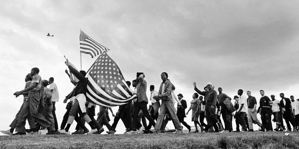 Black and white photo of a large group of people walking on a dirt road from right to left. Two people to the left side of the image are holding American flags.