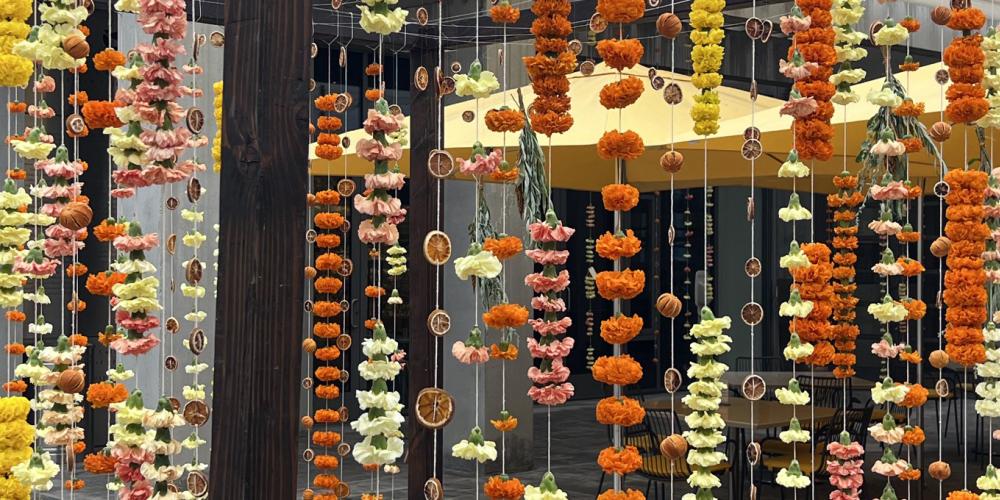 Strings of yellow and orange carnations and mums, and orange slices hung vertically on a wooden structure creating a cozy shelter for a table and chairs.