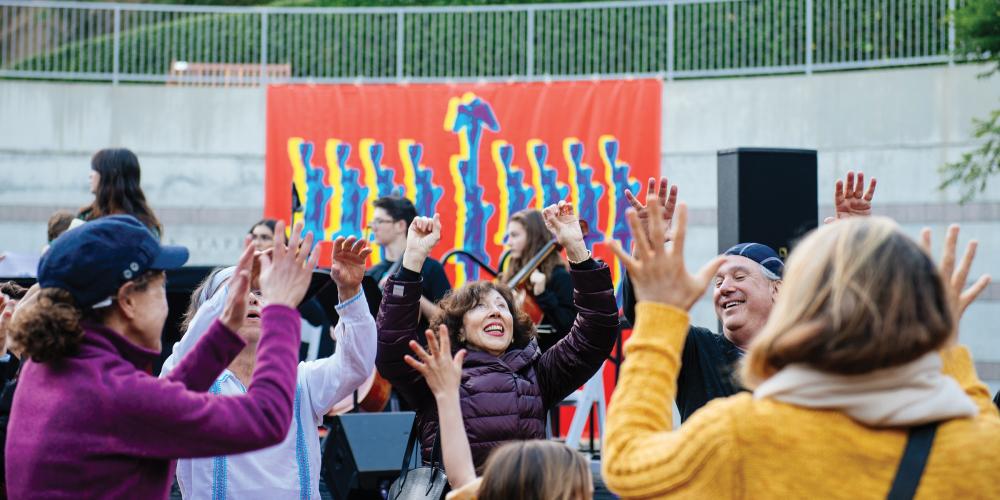 A group of people dancing outside in front of a large banner of a hanukkiah