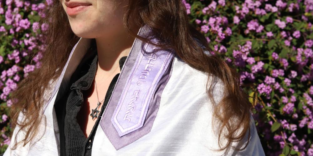 A woman wearing a purple tallit and silver Star of David necklace standing outside in front of a bush of purple flowers.