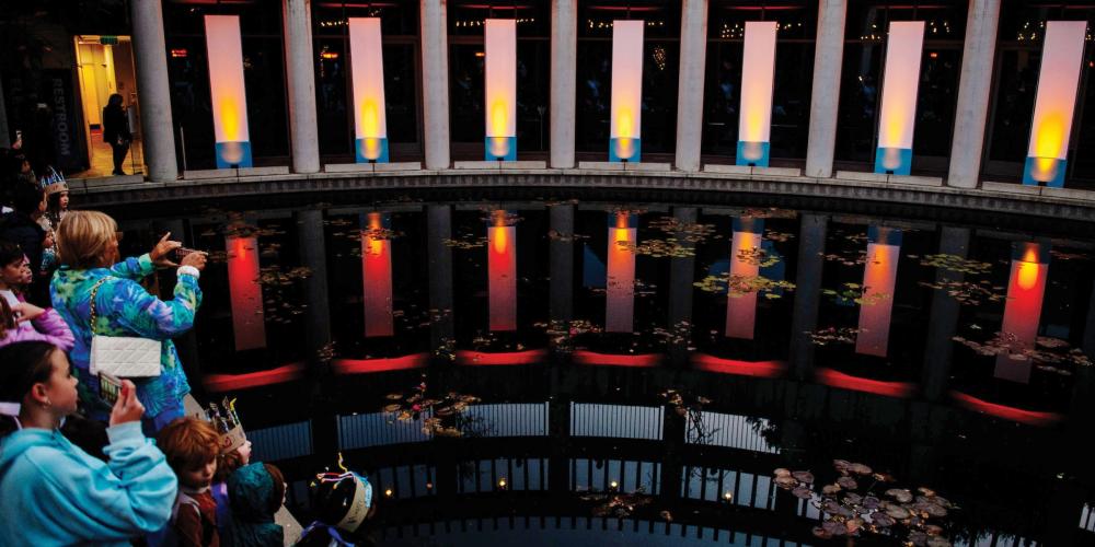 Photo of the lily pond at the Skirball lit up with large column spotlights mimicking a Hanukkah. Families stand along the pond looking at the lights.